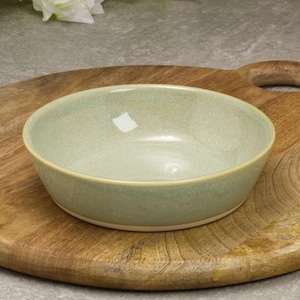 May include: A pale green ceramic bowl with a textured finish, placed on a wooden chopping board. The bowl has a rounded form and a light beige rim. The chopping board features a handle and natural wood grain patterns.