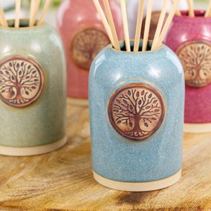 May include: Four ceramic reed diffuser bottles in green, pink, blue, and red. Each bottle features a tree of life emblem and contains wooden reeds. The bottles are on a wooden surface.