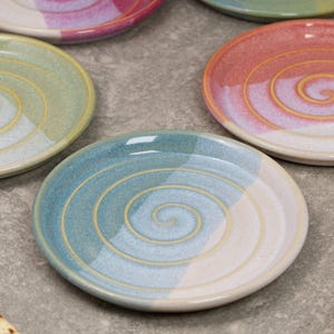May include: A selection of ceramic plates with a spiral pattern. The plates are in various colours, such as blue, green, pink, and white. The plates are round and look handmade.