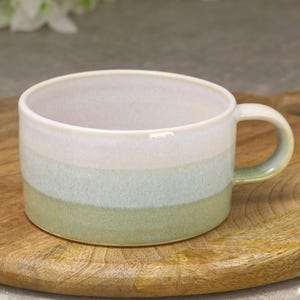 May include: A ceramic mug with a rounded shape and a handle. The mug has a gradient design, transitioning from white at the top to light blue and then to a green base. It sits on a wooden chopping board.