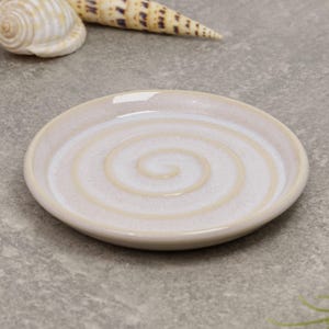 May include: A small, round, off-white ceramic plate with a spiral design in the centre. The plate has a slightly raised edge and a glossy finish. The plate is approximately 10 cm in diameter. Decorative seashells are in the background.
