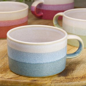 May include: A collection of ceramic mugs with a striped design in blue, white, and green. The mugs have a rounded shape with a handle and are set on a wooden surface. The mugs are suitable for tea, coffee, or other drinks.