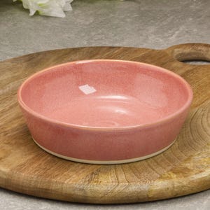 May include: A round, pink ceramic bowl with a white base sits on a wooden cutting board. The bowl has a smooth, glossy finish and a slightly speckled appearance. The cutting board is light brown with a handle.