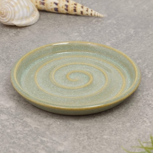 May include: A small, round ceramic dish with a spiral pattern in the centre. The dish is a muted green colour with a golden-brown rim. It is likely used for serving small portions of food or as a decorative item.