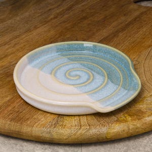 May include: A ceramic spoon rest with a blue and white spiral design. The spoon rest is round with a small lip on one side. The spoon rest is on a wooden cutting board.