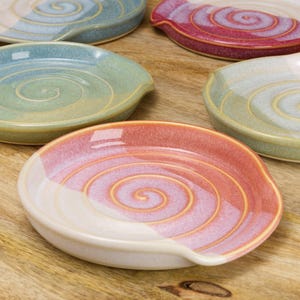 May include: A selection of ceramic spoon rests in shades of blue, green, and red. Each rest has a spiral pattern and a curved rest for a spoon. The spoon rests are presented on a wooden surface.