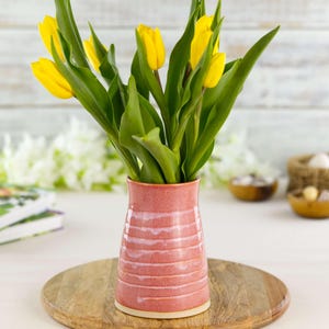 May include: A pink ceramic vase with horizontal white stripes holds a bouquet of yellow tulips with green leaves. The vase sits on a wooden serving board. Other items in the background include books and small bowls.