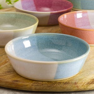 May include: A selection of small, round ceramic bowls in shades of blue, pink, and green. Each bowl features a two-tone design, with a light base and a contrasting interior colour. The bowls are displayed on a wooden surface.