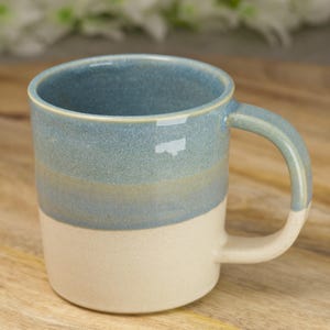 May include: A ceramic mug in light blue and cream. The mug has a rounded handle and a cylindrical shape. The top section is light blue, fading to cream at the base. The mug is designed for holding hot or cold drinks, such as tea or coffee.
