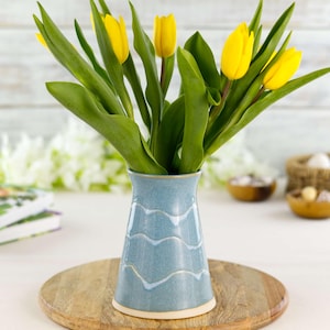 May include: A light blue ceramic vase with a wavy white design holds a bouquet of yellow tulips with green leaves. The vase sits on a wooden round tray. The background includes books and decorative items.