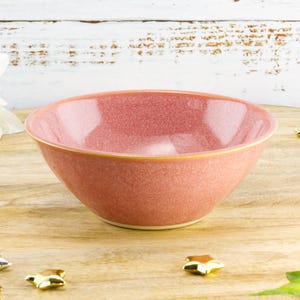 May include: A round, speckled, pink ceramic bowl with a yellow rim. The bowl is set on a wooden surface with star-shaped decorations. The bowl is likely used for serving food.
