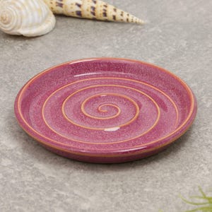 May include: A round, ceramic plate in a deep magenta hue, featuring a golden spiral design. The plate has a slightly raised edge and a glossy finish. Decorative seashells are in the background. This plate is suitable for serving appetizers or as a decorative piece.