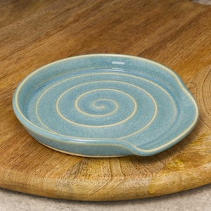 May include: A pale blue ceramic spoon rest featuring a central spiral pattern. The spoon rest is positioned on a wooden chopping board. The spoon rest has a small lip on one side.