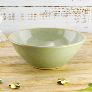 May include: A light green ceramic bowl with a gold rim sits on a wooden surface. The bowl has a rounded shape and a smooth, matte finish. Decorative gold and silver star-shaped objects are scattered around the bowl.