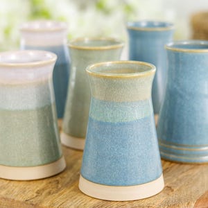 May include: A collection of six ceramic vases in varying shades of blue, green, and cream. The vases have a flared top and a rounded base, with a smooth, glazed finish. They are arranged on a wooden cutting board.