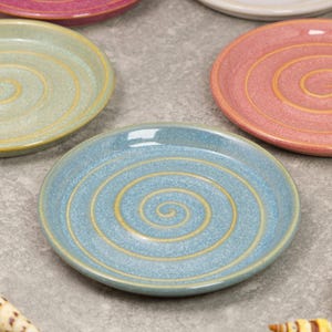 May include: A collection of ceramic plates in various colors, including blue, pink, and green. Each plate features a spiral design in a contrasting color. The plates are round and have a glossy finish, suitable for serving food.
