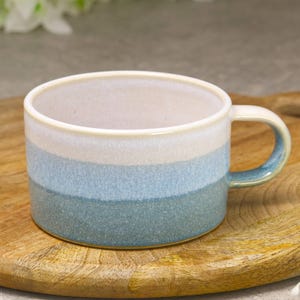 May include: A ceramic mug with a handle, featuring a gradient of colors from off-white to light blue and teal. The mug is set on a wooden cutting board, showcasing its handcrafted appearance. The mug is designed for hot beverages.