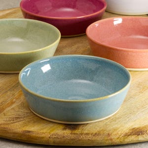 May include: A selection of colourful ceramic bowls in blue, green, pink, and burgundy. The bowls are round with a slightly flared rim and are placed on a wooden serving board. Ideal for serving snacks or small portions.