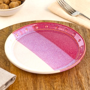May include: A round ceramic plate featuring a two-tone design with a cream-colored section and a vibrant magenta section. The rim is decorated with a series of small, evenly spaced circular indentations that follow the curve of the plate edge.