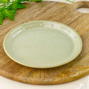 Peut inclure: Une assiette en céramique vert clair avec une surface texturée et une bordure à pois. L'assiette est ronde et repose sur une planche à découper en bois. L'assiette mesure environ 20 cm de diamètre.