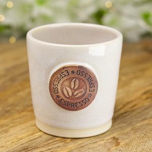 May include: Off-white ceramic espresso cup with a brown, circular emblem. The emblem features three coffee beans and the word "ESPRESSO" in a circle. The cup is on a wooden surface.