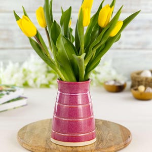 May include: A vibrant magenta ceramic vase with horizontal etched lines holds a bouquet of bright yellow tulips with green leaves. The vase sits on a wooden round tray. The background includes white flowers, books, and small wooden bowls.