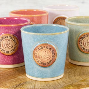 May include: A collection of five ceramic espresso cups in various colours including blue, pink, orange, green, and white. Each cup features a circular brown emblem on the front with the text ESPRESSO and an illustration of three coffee beans.