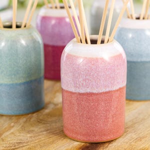 May include: A collection of four cylindrical ceramic reed diffusers, each featuring a two-tone glazed finish in pastel shades of pink, blue, and green. Each vessel contains several light-coloured wooden reeds extending from the open top.