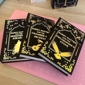 Op de afbeelding: Drie zwarte hardcover boeken met gouden letters en accenten. De titels "Harry Potter and the Prisoner of Azkaban", "Harry Potter and the Chamber of Secrets" en "Harry Potter and the Philosopher's Stone" zijn zichtbaar. De boeken liggen op een roze snijmat.