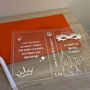 Peut inclure: Couverture de livre en acrylique transparent avec texte et graphiques blancs. La couverture présente le texte "I AM THE ROCK AGAINST WHICH THE SURF CRASHES. NOTHING CAN BREAK ME." et "A COURT OF SILVER FLAMES." Un stylet blanc repose sur la table.
