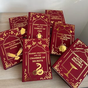 Könnte beinhalten: Eine Sammlung von Harry Potter Büchern mit roten Einbänden und goldener Schrift. Titel sind u.a. "Harry Potter und der Stein der Weisen", "Kammer des Schreckens", "Der Feuerkelch". Goldene Verzierungen umrahmen die Titel.