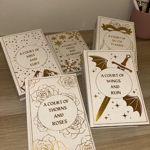 May include: A collection of five white books with gold foil lettering and accents. The titles include "A Court of Mist and Fury," "A Court of Frost and Starlight," "A Court of Silver Flames," "A Court of Thorns and Roses," and "A Court of Wings and Ruin."