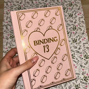 May include: Pink notebook with gold accents and a heart-shaped design that reads "BINDING 13." The cover features a repeating pattern of footballs. The notebook is held over a floral patterned background.