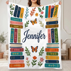 Puede incluir: Una manta blanca de forro polar con una colorida ilustración de libros apilados a ambos lados. El centro muestra el nombre Jennifer en letra cursiva azul oscuro, rodeado de mariposas naranjas, hojas verdes y pequeñas flores.