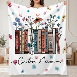 May include: White throw blanket featuring a design of colorful books with floral accents and butterflies. The books have the text "CUSTOM TEXT" on their spines. The blanket also has the words "Custom Name" in a script font.