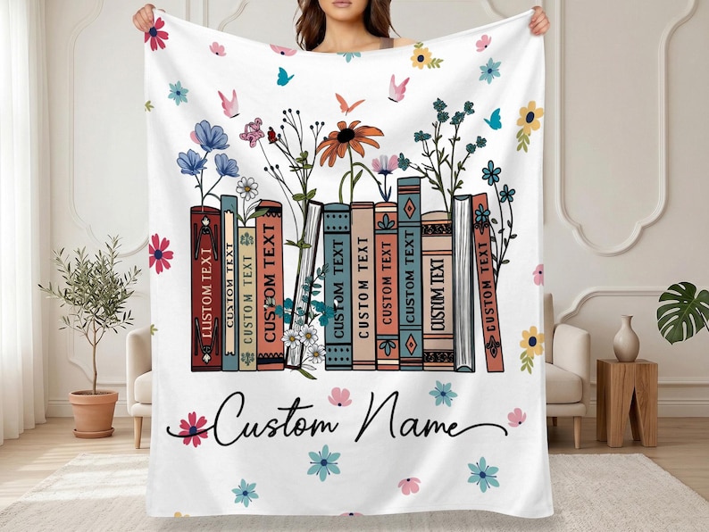 May include: White throw blanket featuring a design of colorful books with floral accents and butterflies. The books have the text "CUSTOM TEXT" on their spines. The blanket also has the words "Custom Name" in a script font.