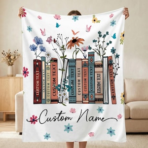 May include: White blanket featuring a floral and book design. The design includes colorful flowers, butterflies, and a row of books with the text "CUSTOM TEXT". The blanket also has the text "Custom Name".