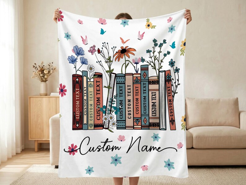 May include: White blanket featuring a floral and book design. The design includes colorful flowers, butterflies, and a row of books with the text "CUSTOM TEXT". The blanket also has the text "Custom Name".
