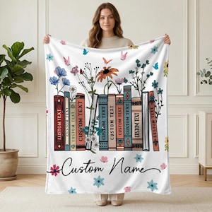 May include: White blanket featuring a floral and book design. The design includes colorful flowers, butterflies, and a row of books with the text "CUSTOM TEXT". The blanket also has the words "Custom Name" in a script font.