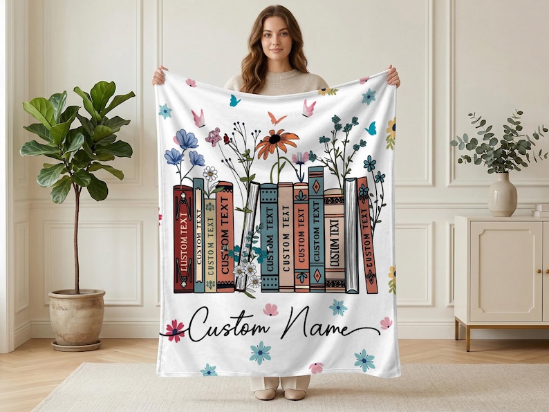 May include: White blanket featuring a floral and book design. The design includes colorful flowers, butterflies, and a row of books with the text "CUSTOM TEXT". The blanket also has the words "Custom Name" in a script font.
