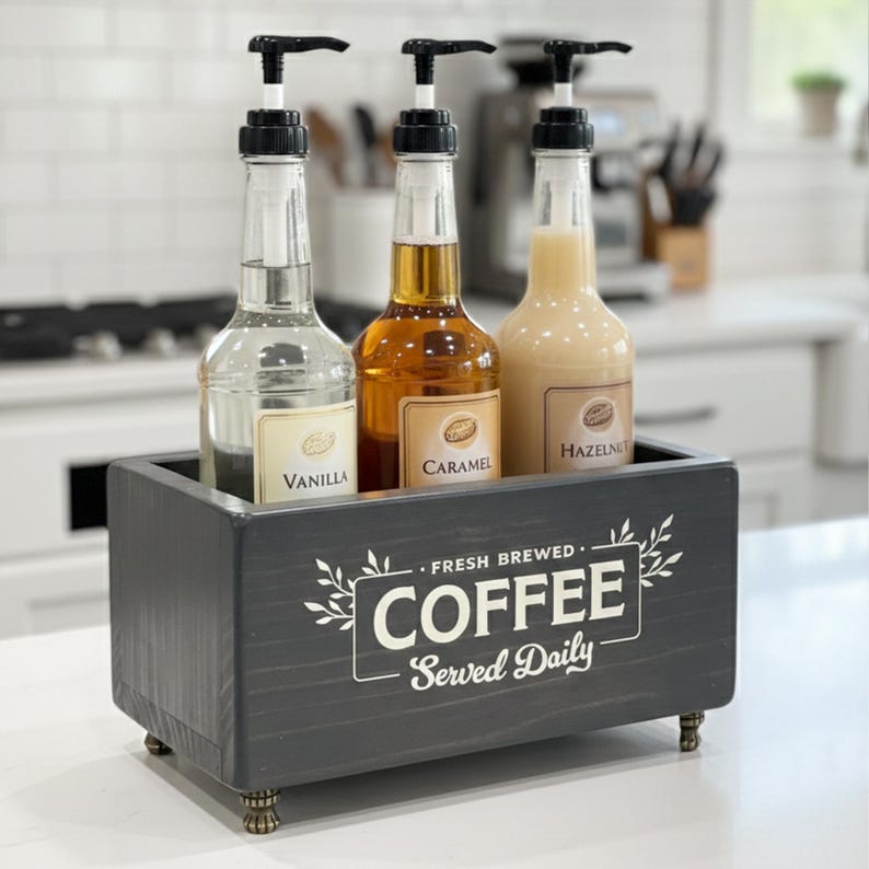 May include: A dark gray rectangular wooden box with four small feet, featuring the white text "FRESH BREWED COFFEE Served Daily". Inside are three clear plastic bottles with black pump dispensers, labeled Vanilla, Caramel, and Hazelnut syrup.