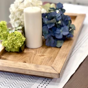 May include: A wooden tray with a natural finish, featuring a white pillar candle and blue and white hydrangea flowers.