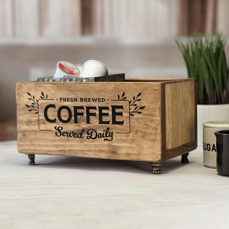 May include: A rectangular wooden storage box with four small metal feet. The front panel features black text that reads FRESH BREWED COFFEE Served Daily, framed by decorative leaf illustrations. The box contains several white coffee pods.