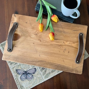 May include: A rectangular wooden serving tray with two leather handles. The tray has a natural wood finish and is decorated with a subtle geometric pattern. The tray is sitting on a green and brown patterned surface.