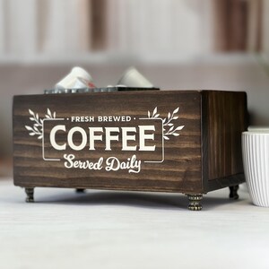 May include: A rectangular dark brown wooden storage box with four small metallic feet. The front features white printed text that reads FRESH BREWED COFFEE Served Daily, framed by a simple rectangular border with decorative leaf accents on the sides.