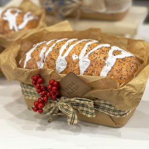 May include: A loaf of bread with white icing and poppy seeds, presented in a brown paper-lined basket. The basket is decorated with a red berry sprig and a plaid ribbon. A tag that reads "FRESH BAKED" is attached to the basket.