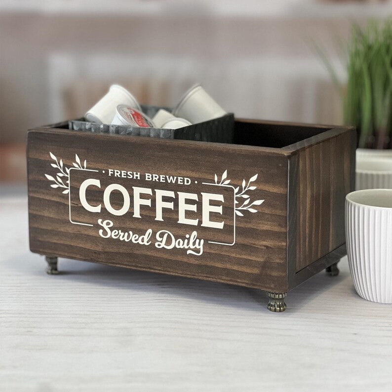 May include: A rectangular dark brown wooden storage box with four small feet. The front features white text that reads FRESH BREWED COFFEE Served Daily, framed by leaf illustrations. Inside the box sits a dark gray metal container holding coffee pods.