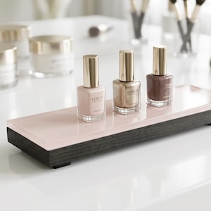 May include: Three glass nail polish bottles with gold caps, labeled Luxury, in shades of pale pink, metallic gold, and mauve, arranged on a rectangular display stand with a glossy pale pink top and a dark charcoal wooden base.