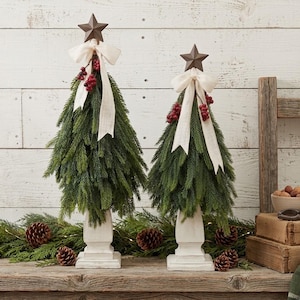 May include: Two artificial evergreen trees on white square pedestals, each topped with a brown metal star. The trees are decorated with cream-colored fabric bows and clusters of red berries, featuring realistic green pine needles and branches.