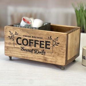 May include: A rectangular wooden storage box with four small metal feet. The front features black text that reads FRESH BREWED COFFEE Served Daily, framed by leaf illustrations. Inside, a metal container holds several white coffee pods.